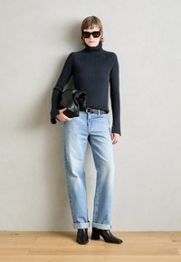 Maglione a collo alto a coste nere, jeans a coste blu chiaro arrotolati, stivaletti neri con tacco, che tiene una pochette nera contro uno sfondo texturizzato.