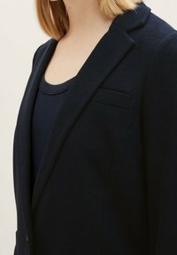 Námořnický blazer vyrobený z texturované látky, s klopovým límcem, náprsní kapsou a vypasovaným střihem, doplněný o odpovídající top.