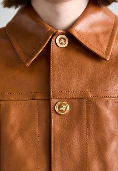 Braune Lederjacke mit großem Kragen, ausgestattet mit goldenen, mit Löwenprägung verzierten Knöpfen und sichtbaren Nähdetails. Glatte Textur, maßgeschneiderter Schnitt.