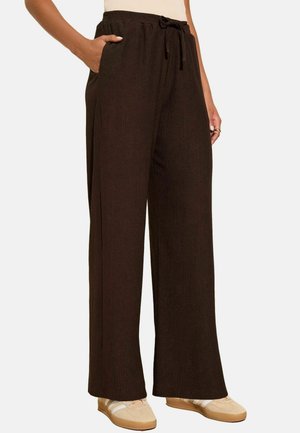 Femme portant un pantalon large marron foncé avec une taille à cordon de serrage et des baskets décontractées beiges, se tenant avec une main dans sa poche.