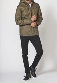 Mężczyzna w oliwkowej kurtce z kapturem, czarnych slim-fit spodniach i czarnych trampkach z białymi podeszwami, stojący na jednolitym tle.