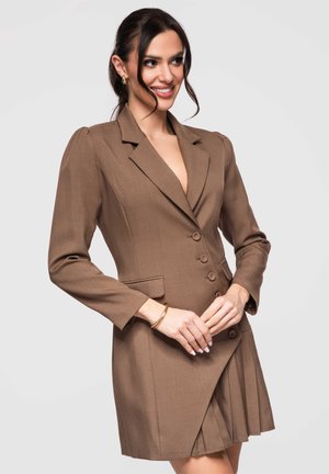 Kobieta w brązowej, dopasowanej sukience marynarkowej z guzikami, złotymi kolczykami kołami i bransoletką, uśmiechająca się i patrząca w bok.