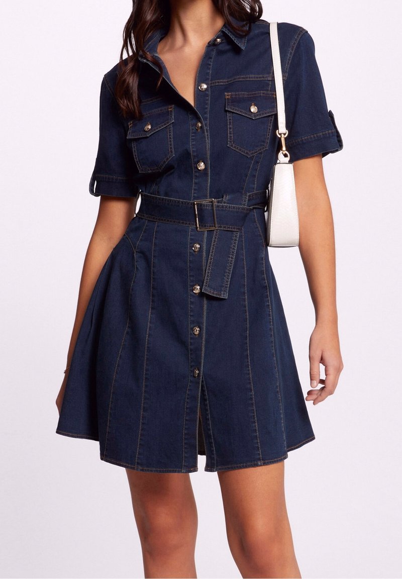 Femme portant une robe en denim bleu foncé à manches courtes, ceinturée, avec des boutons à l'avant et des poches poitrine à rabat, portant un petit sac à bandoulière blanc.