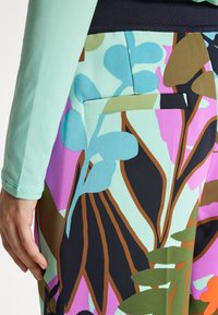 Gros plan d'une main à côté d'une poche de jupe colorée présentant des motifs floraux abstraits aux tons bleus, verts, marron, noir et violet.