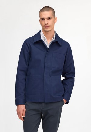 Mężczyzna z krótkimi włosami, ubrany w granatową kurtkę na białą koszulę z kołnierzykiem i ciemne spodnie, stojący na jednolitym jasnym tle.