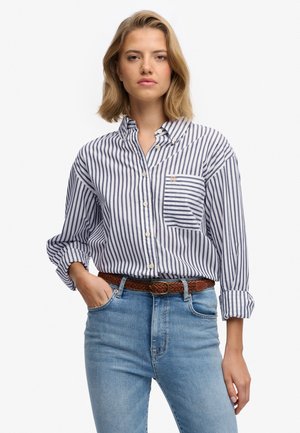 Kobieta o blond włosach do ramion, ubrana w biało-granatową koszulę w pionowe paski, jasnoniebieskie dżinsy i brązowy pleciony pasek.