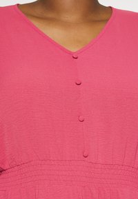 Rosa klänning med V-ringning, fyra knappproppar och en rynkad midja för en avslappnad passform; tillverkad av mjukt, strukturerat material.