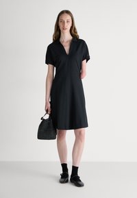 Robe noire à manches courtes avec un col, un décolleté en V et une coupe droite. Le mannequin tient un petit sac à main tissé noir, associé à des chaussettes et des chaussures noires.