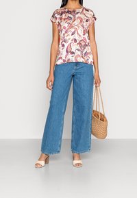 Trykt bluse med korte ærmer i creme, med et flerfarvet paisleymønster. Kombineret med højtaljede blå jeans med vide ben og beige sandaler med blokhæl.