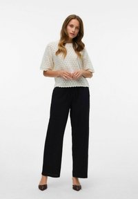Top en tricot ivoire à manches courtes bouffantes, associé à un pantalon noir ample. Le modèle se tient devant un fond neutre, mettant en valeur la tenue.