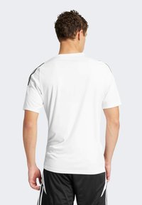 T-shirt de sport blanc à manches courtes, avec des rayures noires le long des épaules. Texture lisse et design ajusté.