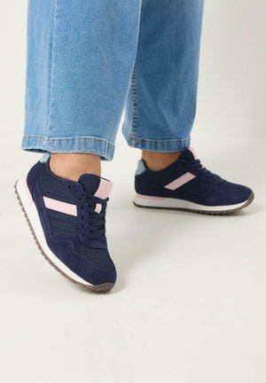 Person iført blå jeans med marineblå og pink atletik-sneakers, der står på et lyst gulv.