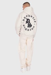 Muž oblečený v smotanových cargo nohaviciach a smotanovej mikine s kapucňou, na ktorej chrbte je čierny gotický text "Dropsize Studios", stojí proti jednoduchému pozadiu.