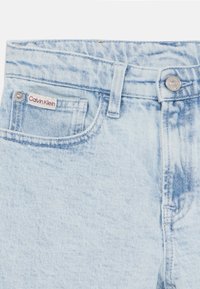 Šviesiai mėlynos džinsinės kelnės su išblukusiu paviršiumi, su priekinėmis kišenėmis, sidabriniu mygtuku ir raudono bei balto "Calvin Klein" etikete.