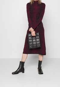Femme portant une robe maroon à motif, arrivant au genou, tenant un sac à main en cuir matelassé noir et portant des bottines en cuir noires.