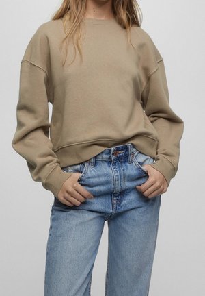 Person iført en løs beige sweatshirt og lyseblå high-waisted jeans med hænderne i forlommerne mod en ensfarvet lys baggrund.