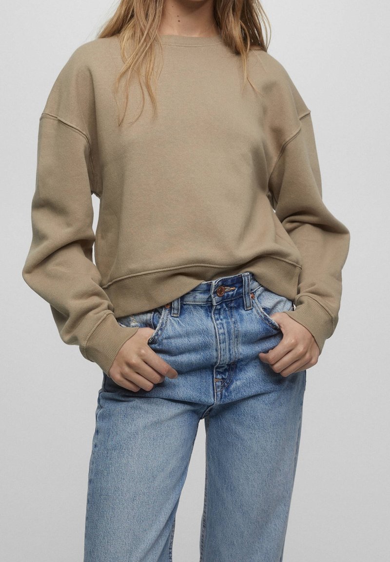 Personne portant un sweat-shirt beige ample et un jean bleu clair taille haute, les mains dans les poches avant, sur un fond clair uni.