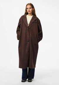 Cappotto in lana marrone con ampi risvolti, bottoni frontali e tasche laterali. Indossato sopra una camicia chiara e pantaloni scuri. Texture liscia.