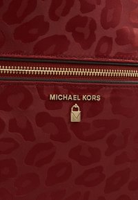 Punainen nailonilaukku, jossa on hillitty leopardikuosi, kultaiset yksityiskohdat ja lukko. "MICHAEL KORS" -logo näkyvissä selvästi.