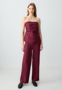 Bordeaux strapless top met gedrapeerde details, gecombineerd met brede broek in dezelfde kleur. Glad linnen textuur, minimale accenten en zwarte schoeisel.