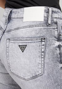 Jeans de denim gris claro con cintura alta, que presentan costuras negras y detalles en los bolsillos. El bolsillo trasero tiene una parche en forma de triángulo negro.