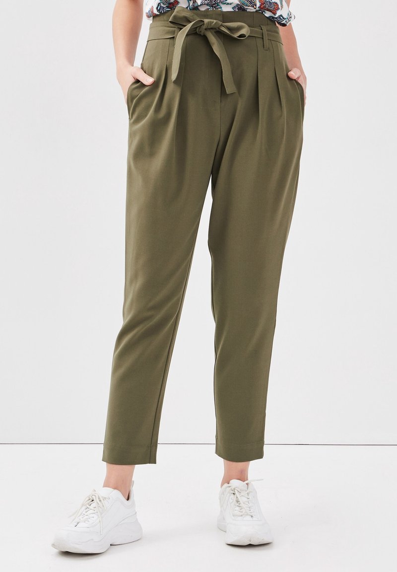 Femme portant un pantalon taille haute olive fuselé avec ceinture nouée, mains dans les poches, associé à des baskets blanches et un haut fleuri.