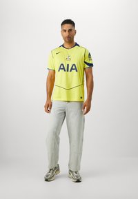 Neon gul fodboldtrøje med blå accenter og stort "AIA" logo, parret med lyse denimbukser og mønstrede sneakers.