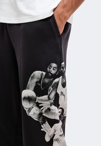 Pantalon noir avec un graphisme blanc représentant un joueur de basket en action tenant un ballon, porté par une personne avec une main dans la poche.