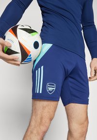 Pantalones cortos de fútbol de color navy con acentos en azul claro, que cuentan con tres franjas y un logo de Arsenal. Se sostiene un balón de fútbol colorido con una mano.