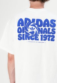Biela bavlnená tričko s odvážnym modrým grafickým textom "ADIDAS ORIGINALS SINCE 1972", so stredným logom a hladkou textúrou.