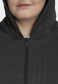 Gros plan d'une personne portant un sweat à capuche Adidas gris foncé avec une fermeture éclair devant et trois bandes verticales sur la poitrine.