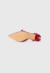Red patent leather high-heeled shoe with a pointed toe and a slim heel. The sole is nude with a textured grip. Adjustable ankle strap detail.