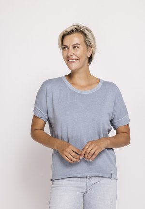 Lächelnde Frau mit kurzen blonden Haaren, die ein hellblaues Kurzarmhemd und hellblaue Jeans trägt, vor einem schlichten weißen Hintergrund stehend.