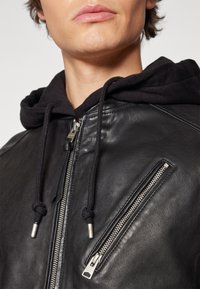 Gros plan d'une personne portant une veste en cuir noire avec des fermetures éclair argentées et un sweat à capuche noir en dessous.