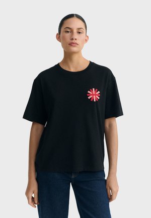 Kobieta ubrana w czarny t-shirt z krótkim rękawem, z małym czerwono-różowym okrągłym wzorem na lewej piersi, w połączeniu z niebieskimi dżinsami.