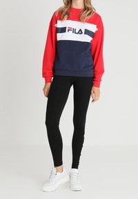 Sudadera de forro polar rojo y azul marino con una franja blanca y el logo de "FILA", combinada con leggings negros y zapatillas blancas.