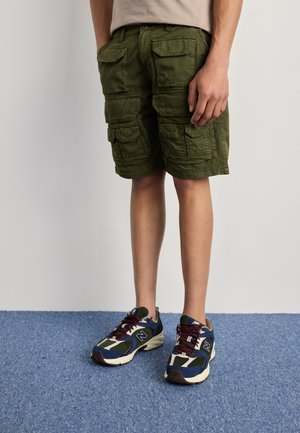 Cargo shorts in olive green, featuring multiple pockets, woven fabric, and a straight cut. Paired with multicolored sneakers and a light shirt.