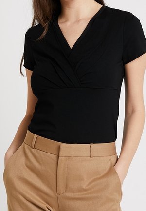 Woman wearing a black short-sleeve V-neck blouse tucked into tan high-waisted trousers with hands in pockets.