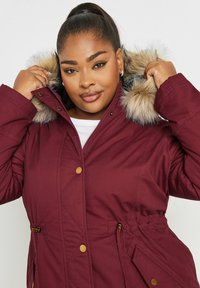 Burgundin väri takki, jossa on tekoturkishuppu, tikattu pinta, kultaiset nappikoristeet ja säädettävä nyöri vyötäröllä.