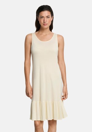 Vrouw in mouwloze, crèmekleurige jurk tot op de knie met gerimpelde zoom, staand tegen een effen lichte achtergrond.