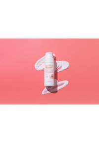 Bottiglia bianca con etichetta "Goodbye Redness Centella Powder Wash", con tappo a ribalta, poggiata su uno sfondo rosa con vortici di polvere bianca.
