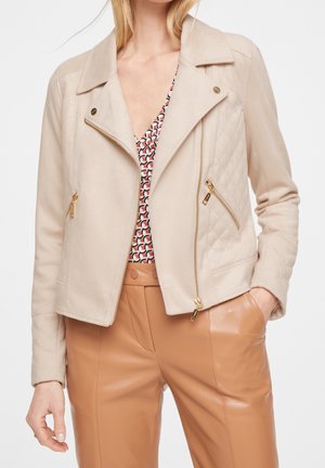 Femme portant une veste matelassée beige avec des fermetures éclair dorées, un haut à col en V à motifs, et un pantalon en cuir beige taille haute, main dans la poche, fond neutre.