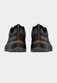 Ζευγάρι πεζοπορικών παπουτσιών Salomon σε σκούρο πράσινο και μαύρο με κίτρινες λεπτομέρειες, από πίσω, σε απλό λευκό φόντο.