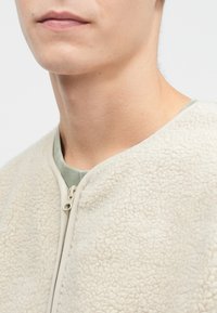 Mikina z fleecu v barvě slonové kosti, s zipovým zapínáním a měkkou strukturou, s lehce zeleným límcem. Detailní pohled.