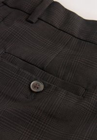 Poche de pantalon sur mesure en tissu à carreaux foncés avec un bouton marron et un passant de ceinture à la taille, montrant la texture et les détails de couture.