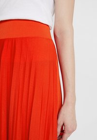 Falda naranja plisada con una alta cinturilla elástica. El material parece ligero y fluido, mostrando una textura suave.