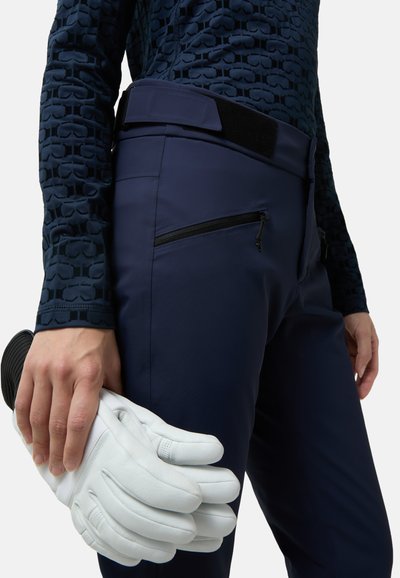 Granatowe dopasowane spodnie z teksturowaną ciemną górą, z zamkiem błyskawicznym w kieszeni. Osoba trzyma białe skórzane rękawiczki w prawej ręce.