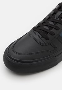 Sneaker in pelle nera con suola in gomma testurizzata. Presenta lacci neri e un sottile accento blu. Superficie liscia con dettagli di cucitura visibili.
