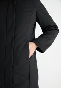 Cappotto nero con una texture liscia, caratterizzato da un colletto alto, una zip nascosta e tasche laterali, mostrato con un braccio appoggiato su una tasca.