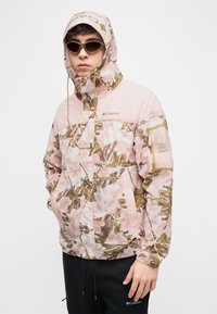 Νέος άνθρωπος που φοράει ροζ ζακέτα με κουκούλα Columbia με λουλουδάτο μοτίβο και μαύρο παντελόνι Columbia, με γυαλιά ηλίου, στέκεται μπροστά σε λευκό φόντο.